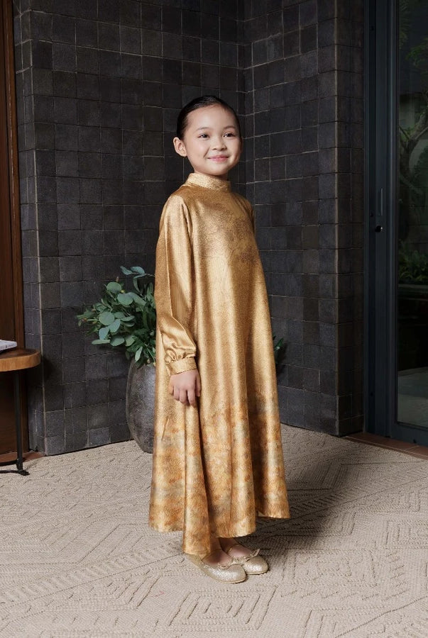 Child wearing a gold dress by Jenna & Kaia standing in a room with a textured wall and a small table.