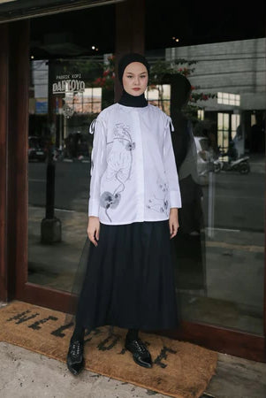A woman wearing a white blouse by Maima with long sleeves and a round neckline, paired with a maxi skirt. The blouse features a floral pattern and she is standing in front of a glass door.