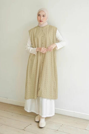 Woman wearing a butter-colored long vest by Mistyvori over a white outfit against a plain white wall.