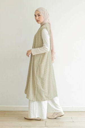 Woman wearing a khaki long vest by Mistyvori over a white shirt with a hijab, standing against a plain white wall.
