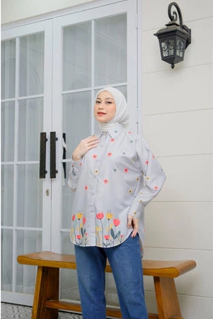 A woman wearing a grey long-sleeved shirt by Abame with a floral pattern, standing on a bench with her hand raised to her face, in front of a door with glass panels and a wall-mounted lamp.