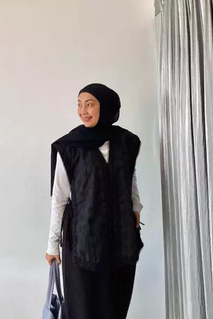 Person wearing a black hijab and black vest by Abame against a white wall with a curtain.