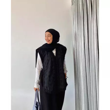 Person wearing a black hijab and black vest by Abame against a white wall with a curtain.