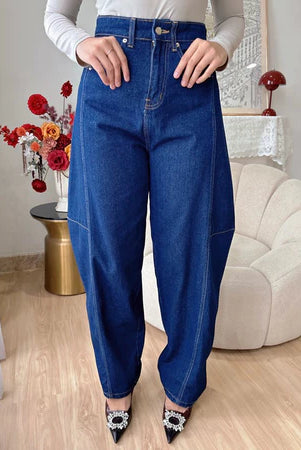 A woman standing in an indoor setting wearing dark blue barrel jeans and white top, with decorative flowers and furniture in the background.
Seorang wanita berdiri di dalam ruangan, mengenakan celana denim model barrel berwarna biru tua dan atasan putih, dengan hiasan bunga dan perabotan di latar belakang.