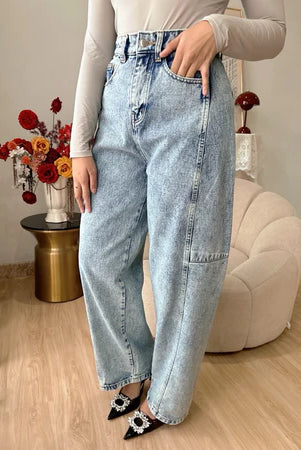 A person standing in an indoor setting wearing light blue washed barrel jeans with a high waist and straight leg from Haidee Orlin, paired with a beige top and black and white shoes.