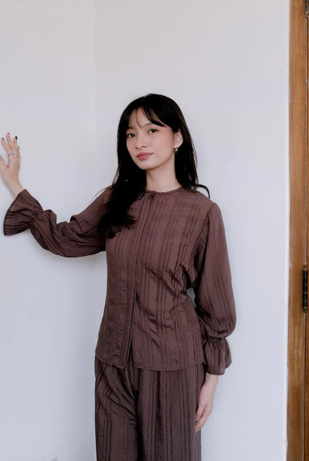 Woman wearing a brown blouse by Renaette against a white wall.