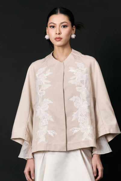 Woman wearing a beige embroidered jacket with white accents by Studio Tui on a black background