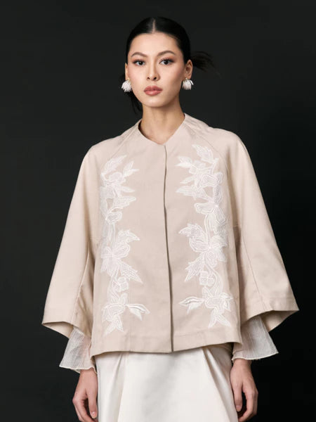 Woman wearing a beige embroidered jacket with white accents by Studio Tui on a black background