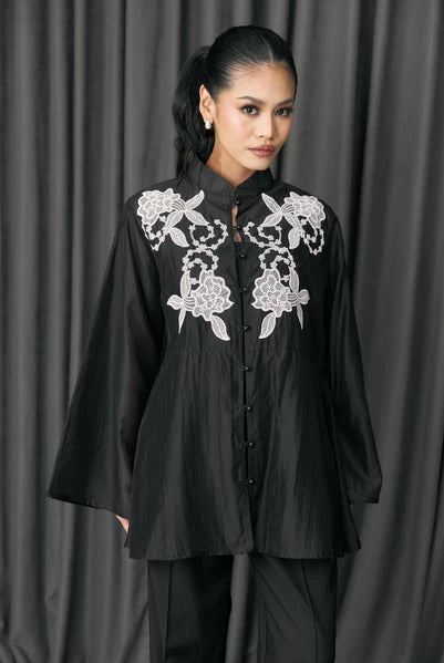 Woman wearing a black blouse with white floral embroidery by Studio Tui against a gray curtain background