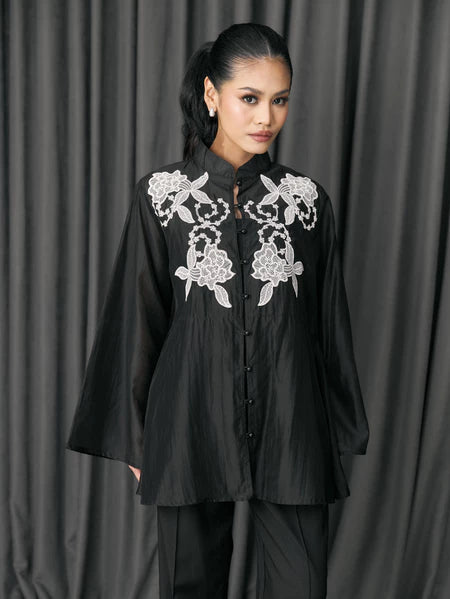 Woman wearing a black blouse with white floral embroidery by Studio Tui against a gray curtain background