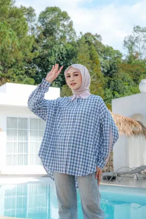 Woman wearing a blue checkered shirt from Abame and hijab by a poolside with trees in the background