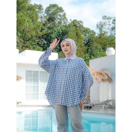 Woman wearing a blue checkered shirt from Abame and hijab by a poolside with trees in the background