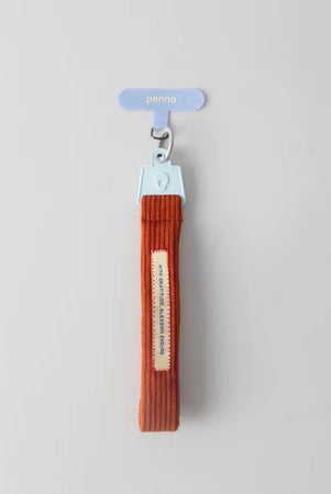 Brick red keychain strap with a metal clip from Muhajir Project on a light gray background