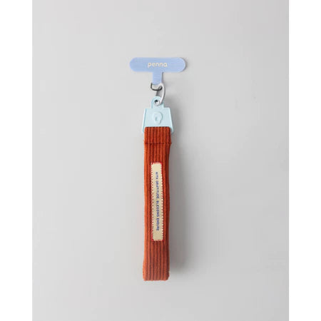 Brick red keychain strap with a metal clip from Muhajir Project on a light gray background