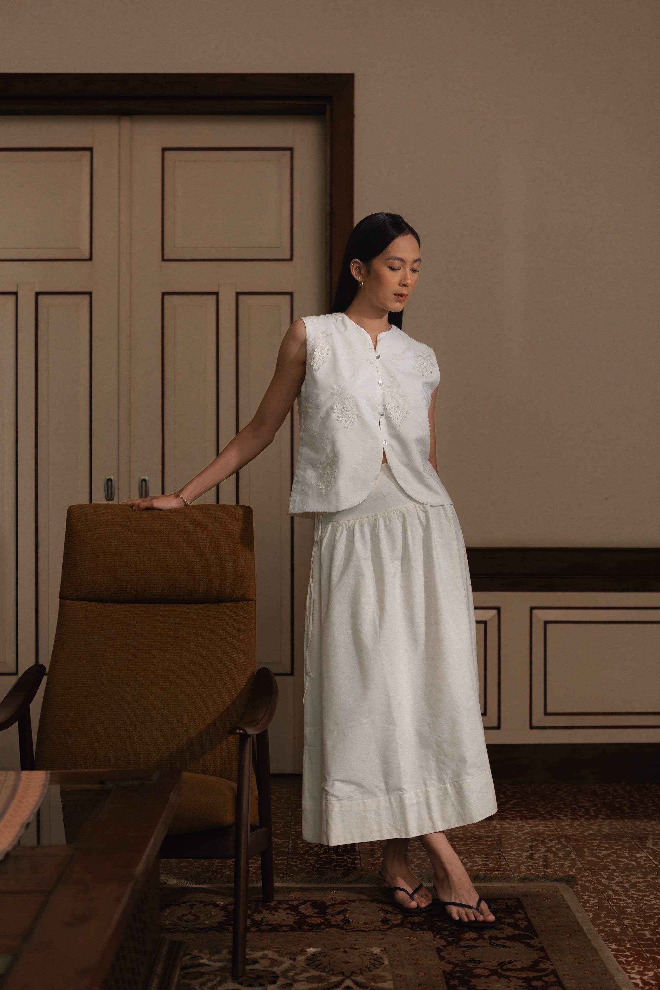 Woman in a white vest by Kasyalia and skirt standing in a room with wooden furniture and a neutral wall.