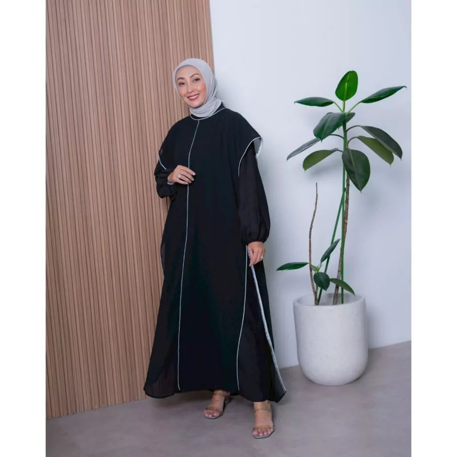 Woman in a black dress by Abame standing next to a plant indoors