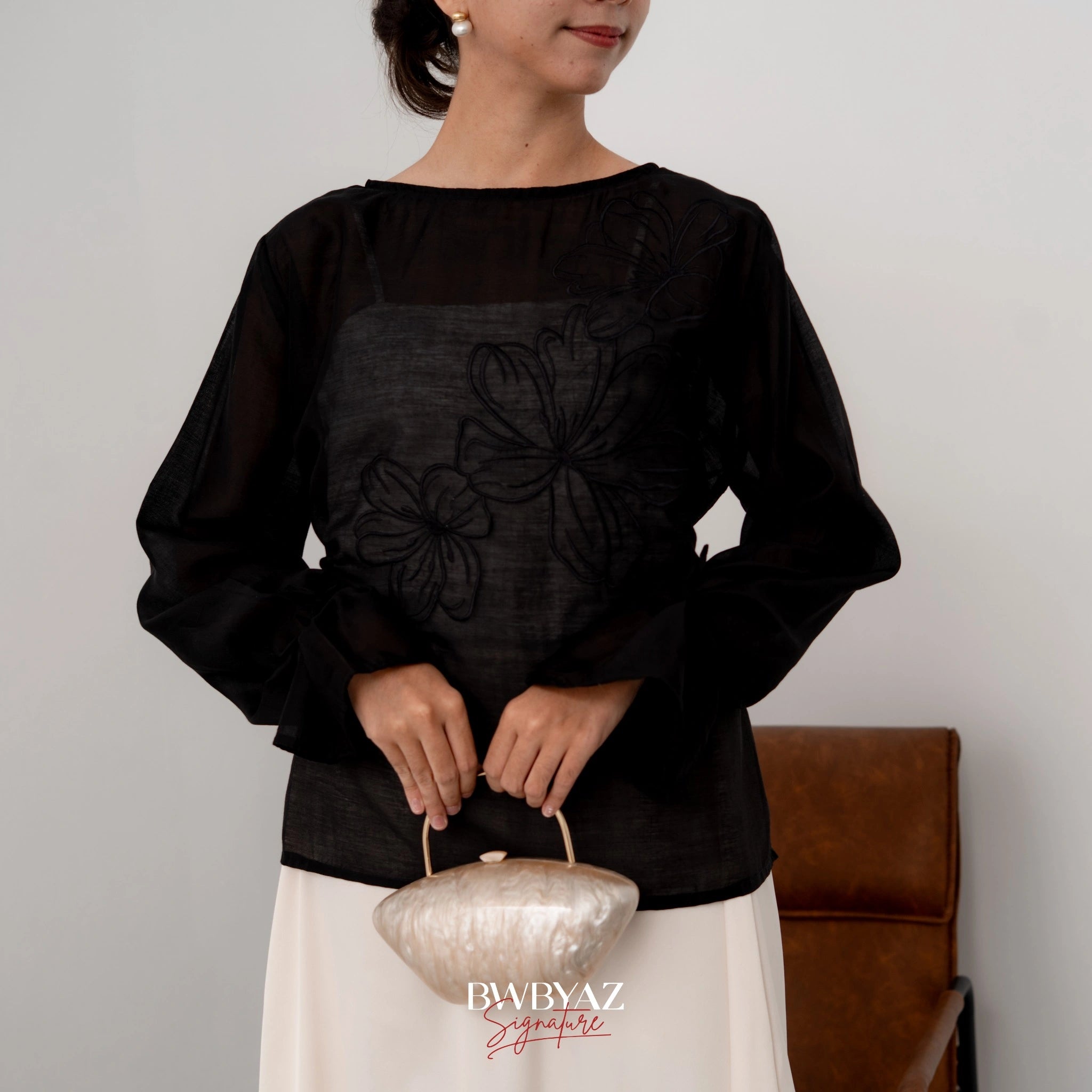 A woman wearing a black blouse with floral embroidery on the front, made from Luminous Sheer Tencel, with tie accents on the sleeves and a small loop at the waist.