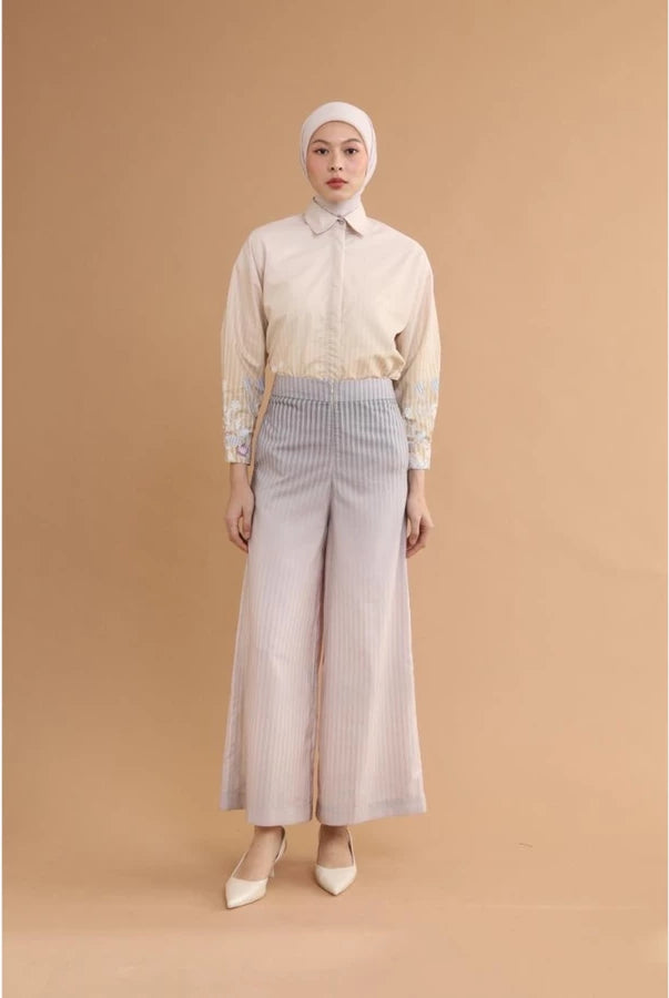 Woman wearing a cream blouse and violet wide-leg pants by Haidee Orlin against a brown background