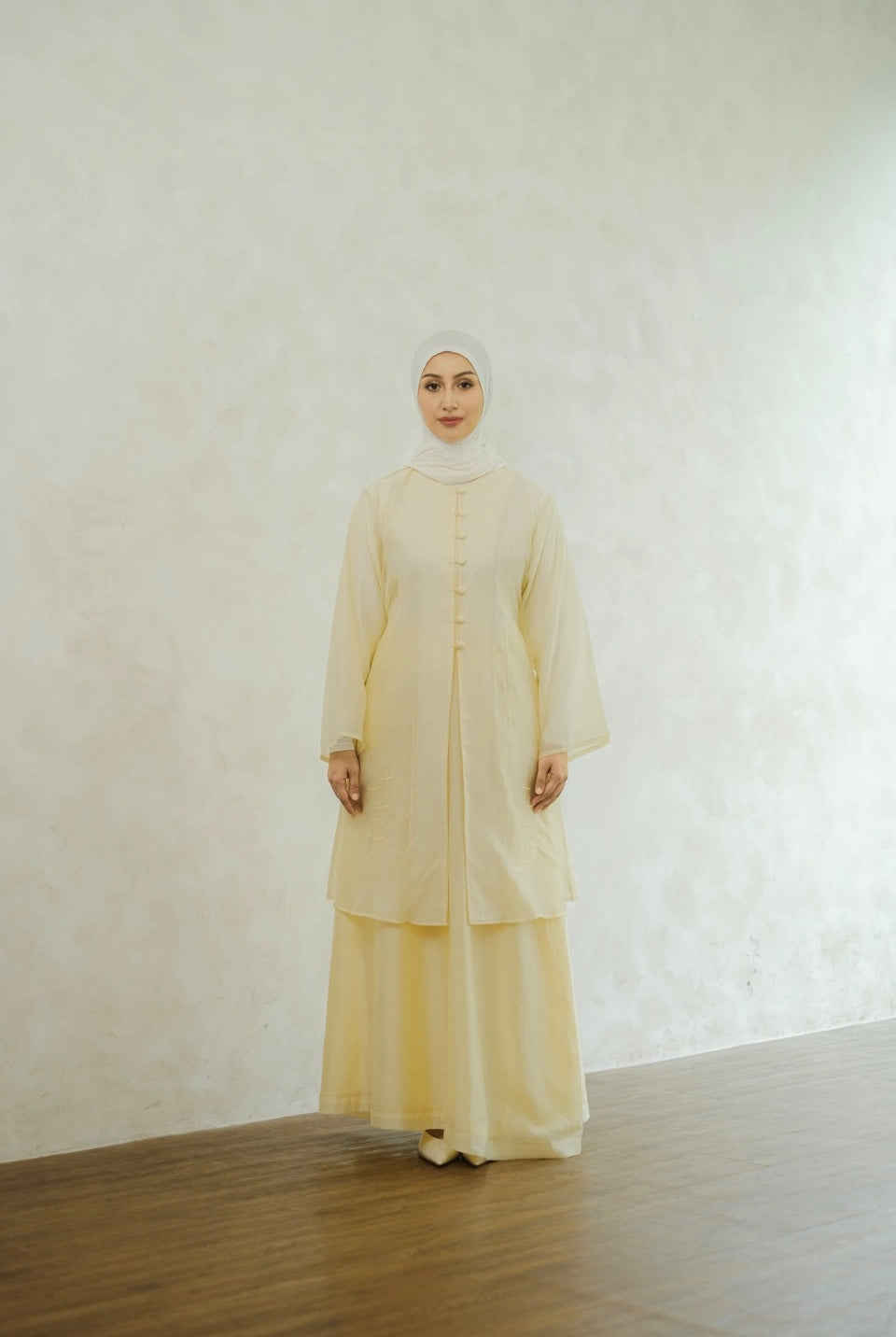 Woman wearing a light yellow tunic by Tufine and hijab against a white wall.
Seorang wanita yang mengenakan tunik berwarna kuning muda dari Tufine dan jilbab, berlatar belakang dinding putih.