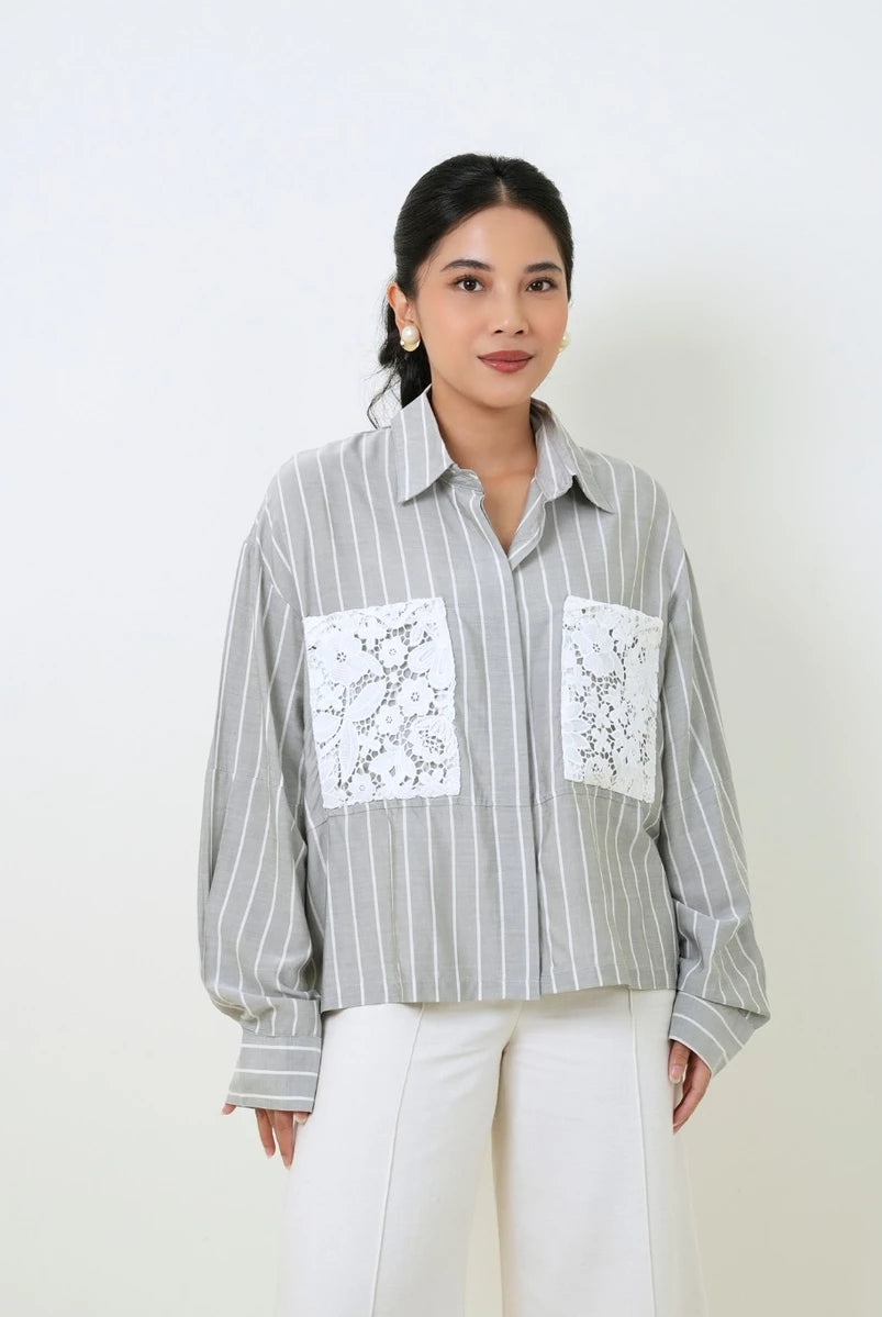 A woman wearing a light gray and white vertically striped button-up shirt by Studio Tui with two white lace pocket flaps, paired with white pants, standing against a plain off-white background.