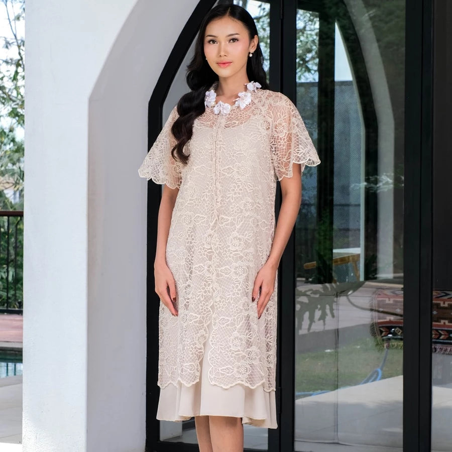 Woman wearing a beige lace outer dress by Holy Peony standing in front of a glass door.