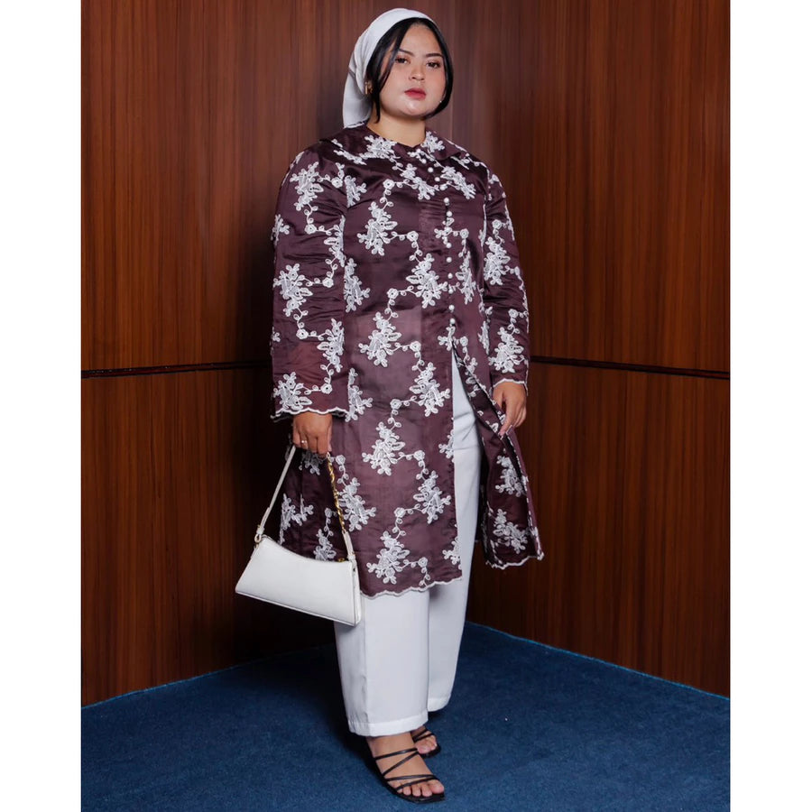 Woman wearing a dark brown color patterned tunic by Vlowy and white pants against a wooden panel background
Seorang wanita mengenakan tunik bermotif berwarna cokelat tua dari Vlowy dan celana putih di depan latar belakang panel kayu