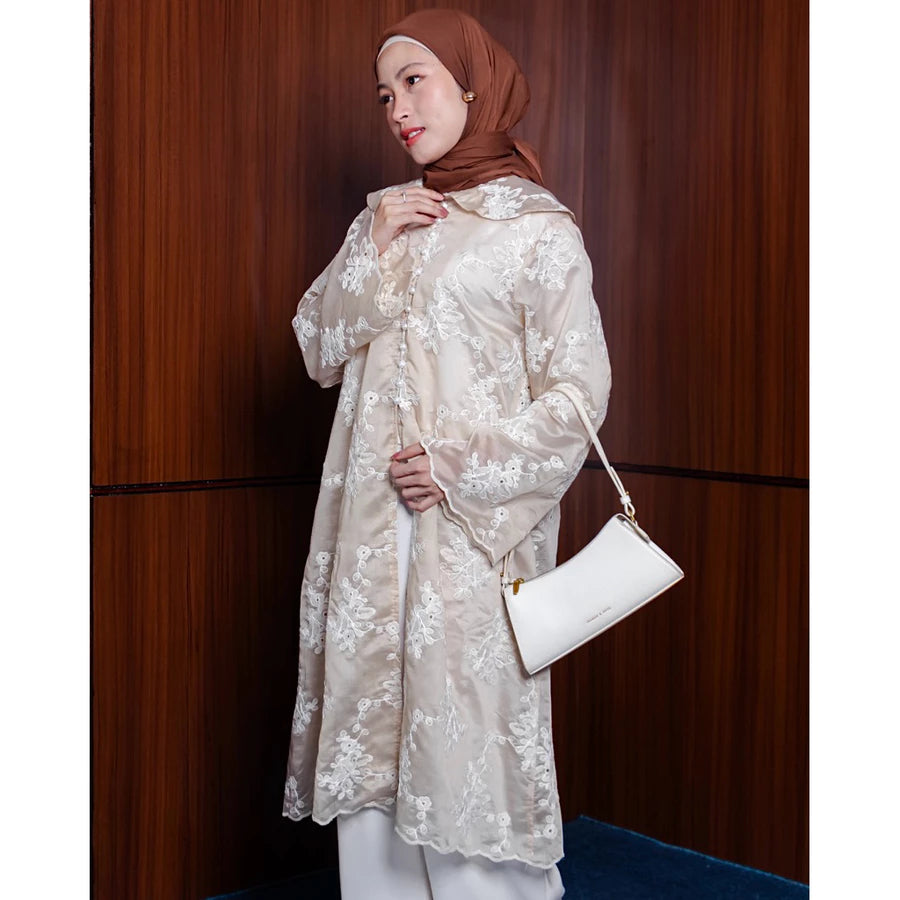 Woman wearing a beige floral lace tunic by Vlowy holding a white handbag against a wooden panel background
Seorang wanita mengenakan tunik bermotif bunga berwarna beige dari Vlowy, memegang tas tangan putih di depan latar belakang panel kayu.