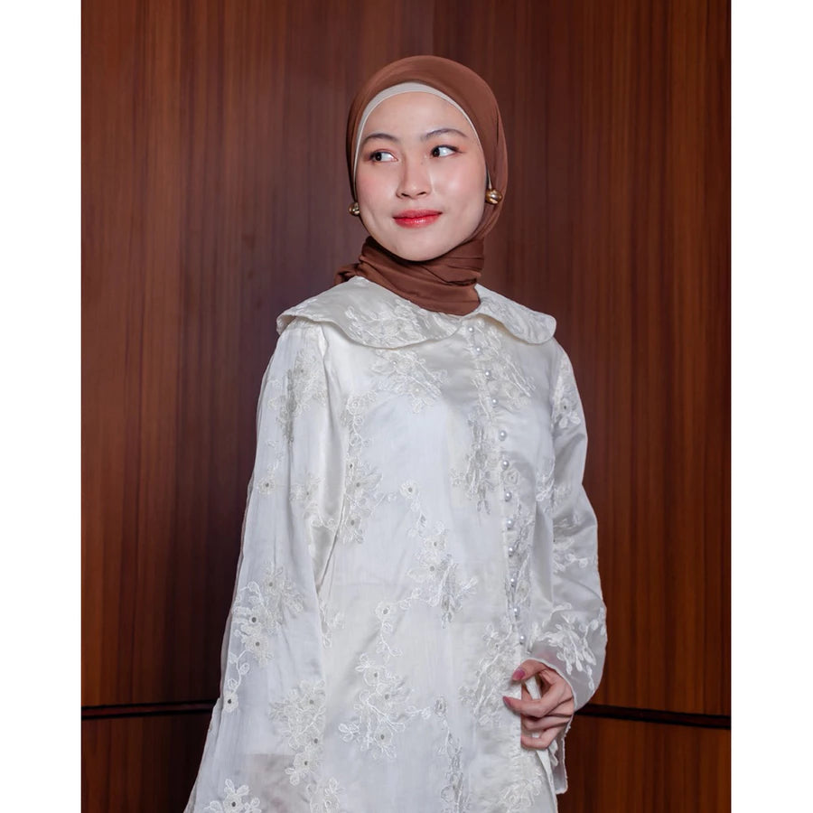 Woman wearing a white lace tunic by Vlowy and brown hijab against a wooden panel background
Seorang wanita mengenakan tunik putih berenda dari Vlowy dan hijab cokelat dengan latar belakang panel kayu.
