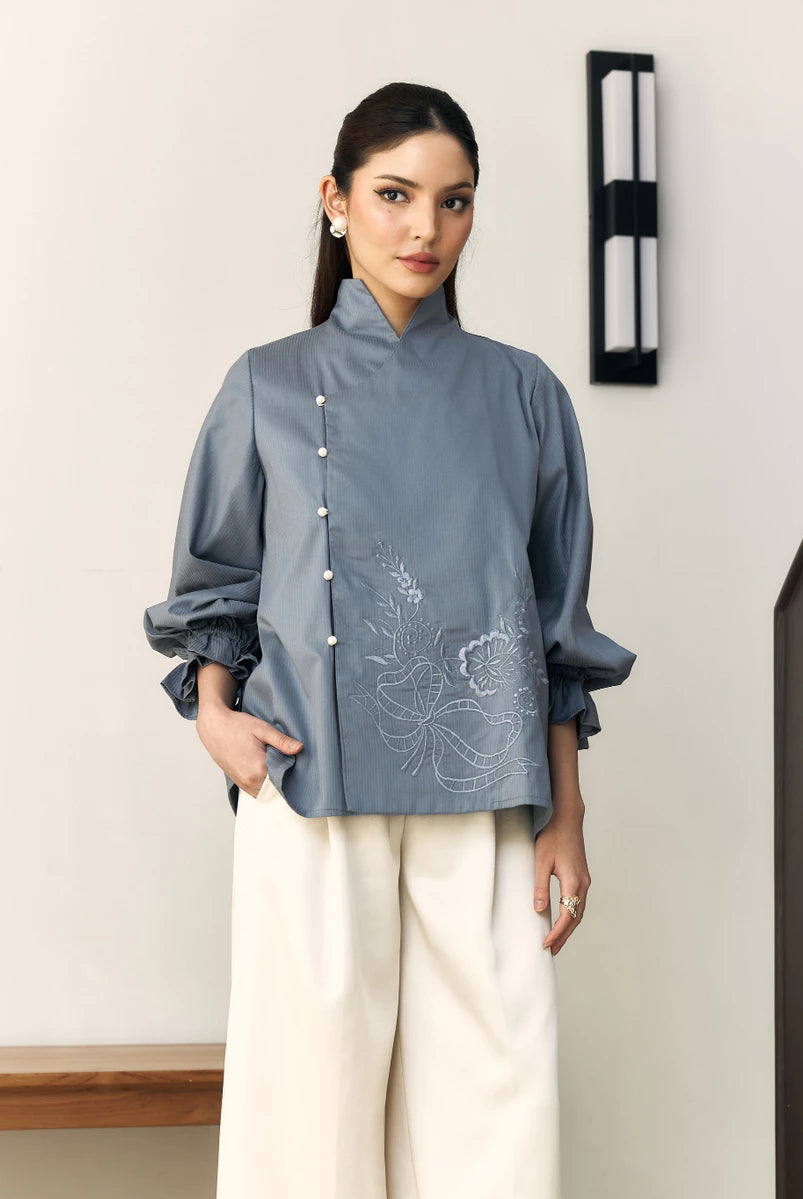 Woman wearing a blue blouse with floral embroidery by Studio Tui and white pants indoors.
