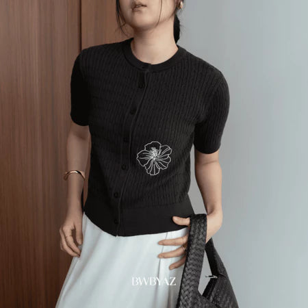 A woman wearing a dark brown short-sleeve cable knit cardigan with a floral embroidery on the front from BWBYAZ, paired with a white skirt.