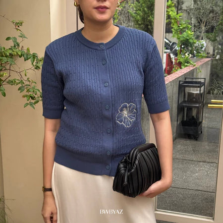 A woman wearing a denim colored short-sleeve cable knit cardigan with a floral embroidery on the front from BWBYAZ, paired with a white skirt.
