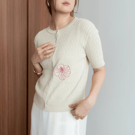 A woman wearing a beige short-sleeve cable knit cardigan with a floral embroidery on the front from BWBYAZ, paired with a white skirt.