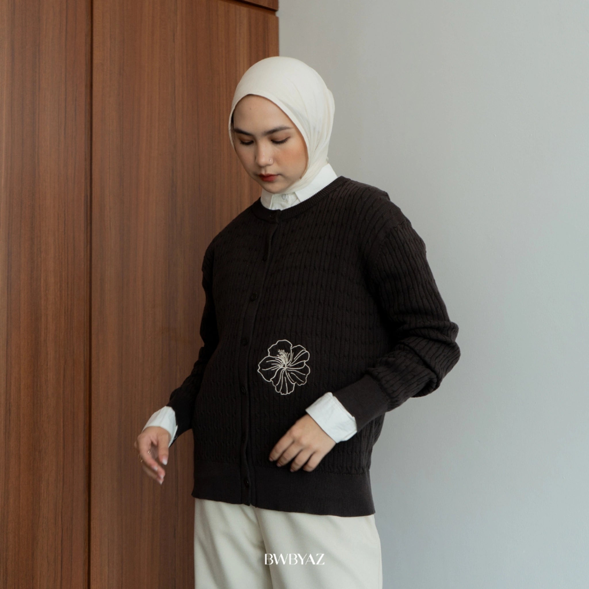Woman wearing a dark brown cable knit long sleeve button-up cardigan with white floral embroidery on the pocket, styled modestly with a cream hijab and ivory trousers.