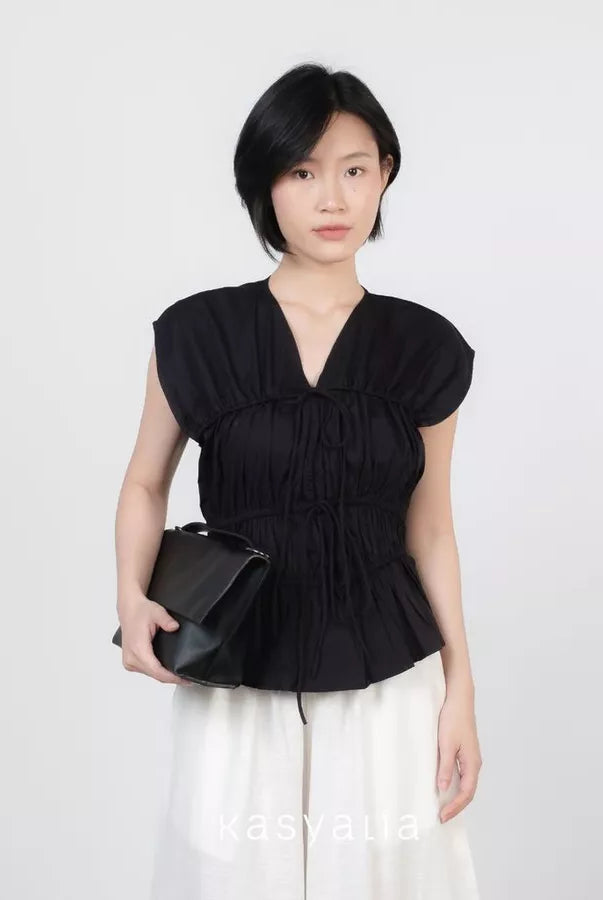 A woman modeling a black sleeveless peplum top by Kasyalia with a gathered V-neckline, paired with white pants and holding a black clutch bag, set against a plain light gray background.