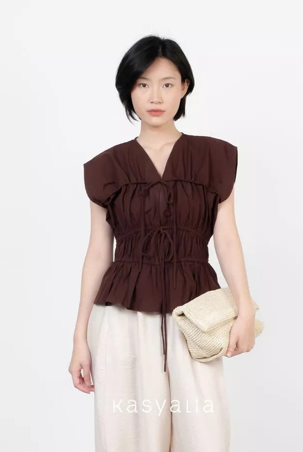 A model wearing a dark brown ruffled peplum top by Kasyalia with a tie waist, paired with cream-colored pants and holding a woven beige clutch bag, set against a plain white background.