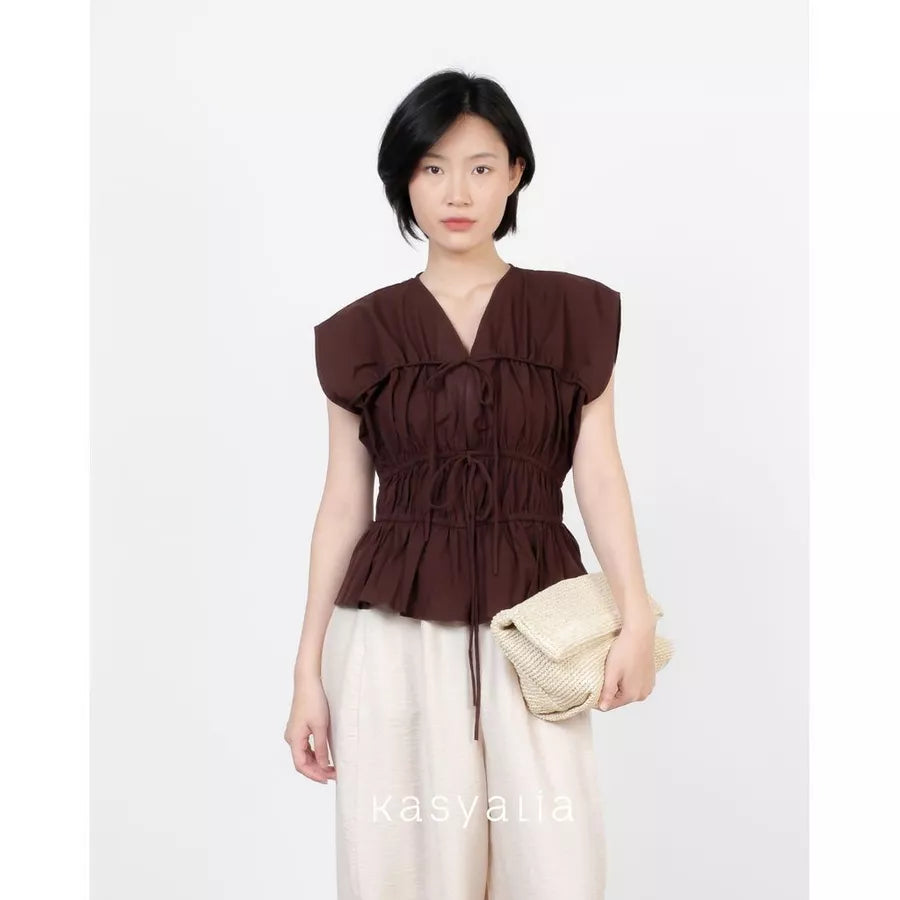 A model wearing a dark brown ruffled peplum top by Kasyalia with a tie waist, paired with cream-colored pants and holding a woven beige clutch bag, set against a plain white background.