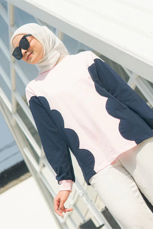 Woman wearing a pink sweater with navy blue patterns by Casa Elana and a hijab, standing on a white railing.