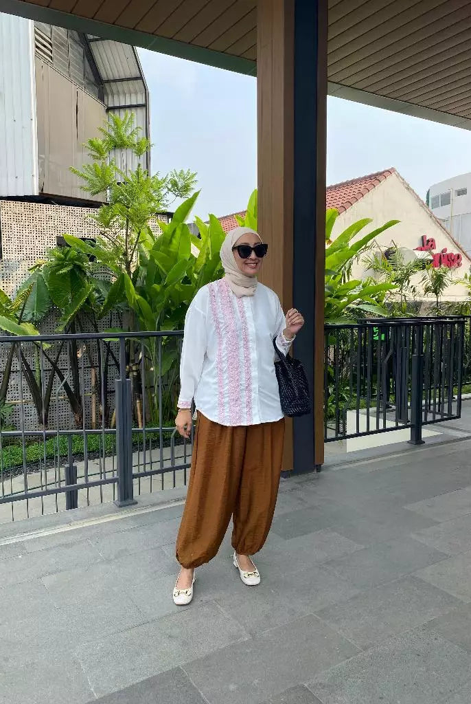Woman in a white top by Abame and brown pants standing outdoors with greenery and buildings in the background