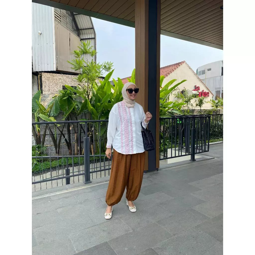 Woman in a white top by Abame and brown pants standing outdoors with greenery and buildings in the background