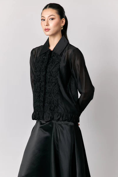 Woman wearing a black sheer shirt by Studio Tui with ruffled details on a plain background