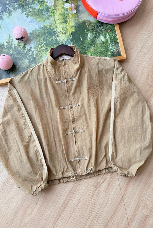 A beige cheongsam jacket by Haidee Orlin with a high collar, laid flat on a light wood floor with decorative props in the background.