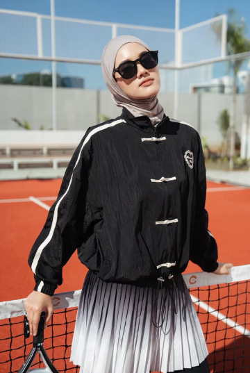 A woman wearing a black lightweight cheongsam jacket by Haidee Orlin with a black-and-white pleated skirt, posing on an outdoor tennis court.
