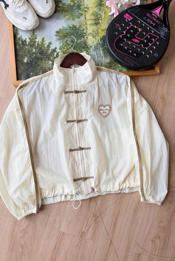 Cream-colored windbreaker cheongsam jacket by Haidee Orlin, laid flat on a wooden floor next to a framed picture, shoes, and a paddle racket.