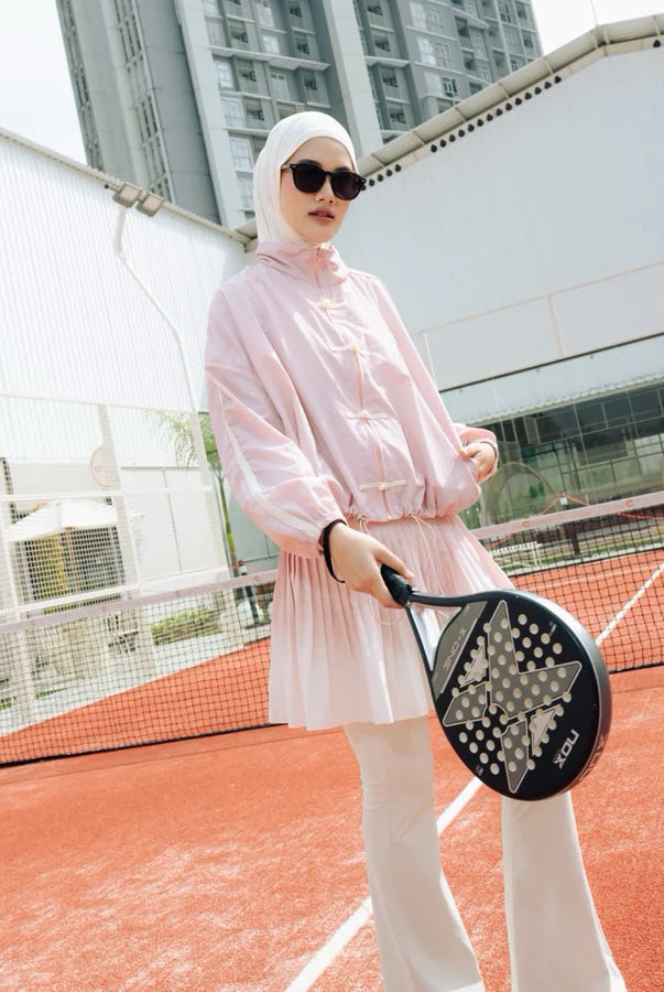 A woman wearing a light pink long-sleeve athletic cheongsam jacket by Haidee Orlin and white pants stands on an outdoor tennis court holding a black paddle racket.