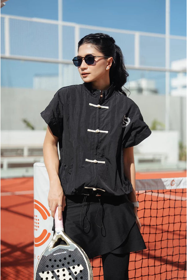 A woman wearing a black cheongsam active vest by Haidee Orlin with black pants stands on an outdoor tennis court holding a paddle racket.