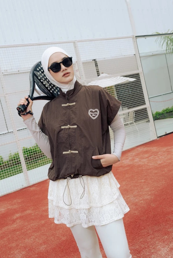 A woman wearing a brown cheongsam vest by Haidee Orlin over a white long-sleeve top and white lace skirt, posing on an outdoor tennis court while holding a racket.