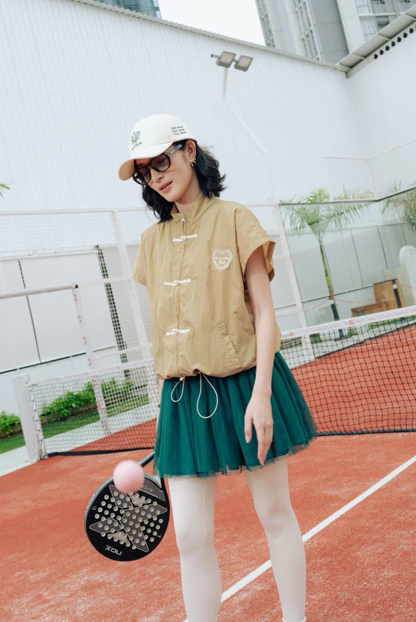 A woman stands on an outdoor tennis court wearing a khaki cheongsam vest by Haidee Orlin with white drawstrings, a dark green pleated tennis skirt, white tights, a white cap, and sunglasses. She holds a paddle with a pink ball.