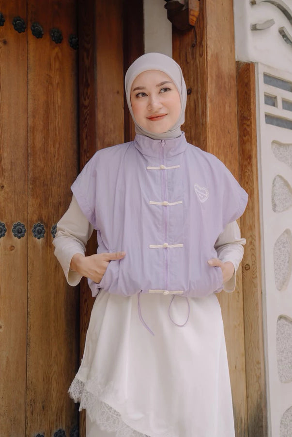 A woman wearing a lilac sleeveless cheongsam vest by Haidee Orlin over a white dress and a white hijab, standing in front of a rustic wooden door.