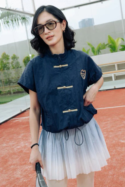 A woman wearing a navy blue cheongsam vest by Haidee Orlin with gold-toned frog closures and a light blue to white ombré tulle skirt, standing on an outdoor tennis court.
