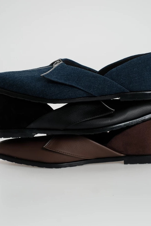 Three pairs of slip-on flat shoes stacked vertically on a light gray background. The top pair is dark navy blue denim with a black sole, the middle pair is black leather with a black sole, and the bottom pair is brown leather with a black sole.